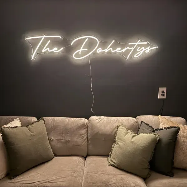 Warm white LED neon script sign reading The Dohertys on a charcoal wall above a taupe sofa with olive cushions